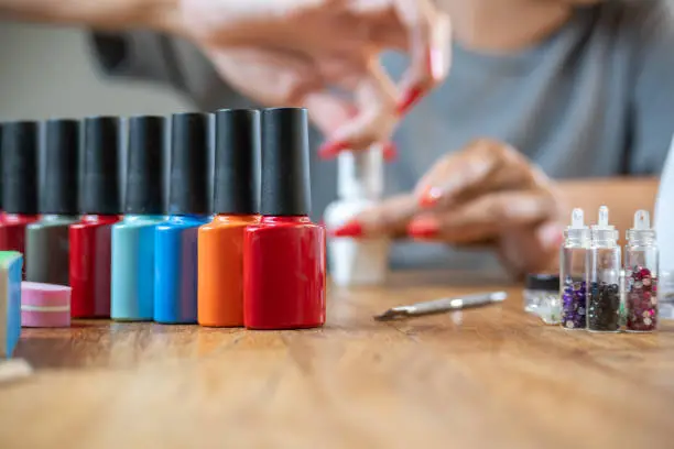 A row of colorful nail polish bottles on a wooden table, with a person doing a manicure blurred in the background. A row of colorful nail polish bottles on a wooden table, with a person doing a manicure blurred in the background.