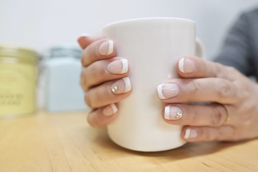 A close-up of a person's hands with a French manicure holding a white ceramic mug on a wooden table. A close-up of a person's hands with a French manicure holding a white ceramic mug on a wooden table.